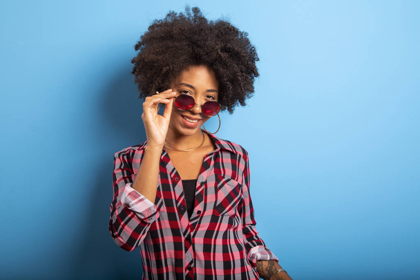 Cheerful attractive afro brazilian girl enjoying life wearing sunglasses On blue wall background - Imagem. - Фото, изображение