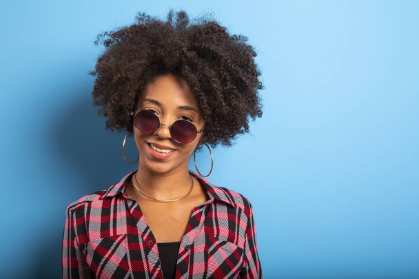 Cheerful attractive afro brazilian girl enjoying life wearing sunglasses On blue wall background - Imagem. - Фото, зображення