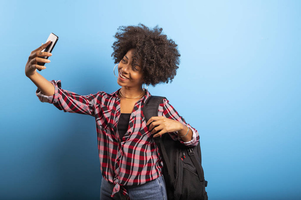 Primer plano retrato de la joven sonriente atractiva mujer brasileña africana sosteniendo teléfono inteligente, tomando foto selfie en el fondo azul
. - Foto, imagen