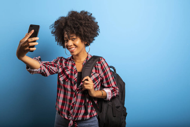 Closeup portrait of smiling young attractive African brazilian woman holding smartphone, taking selfie photo on the blue background. - Foto, afbeelding