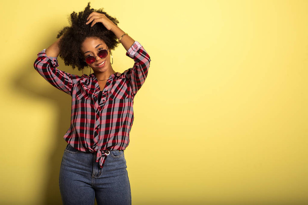 Cheerful attractive afro brazilian girl enjoying life wearing sunglasses On yellow wall background - Imagem. - Foto, immagini