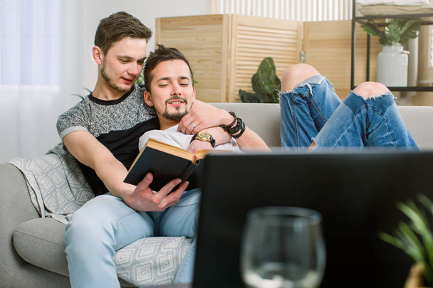 Ελκυστικό γκέι ζευγάρι διαβάζοντας το βιβλίο στον καναπέ στο σπίτι στο καθιστικό - Φωτογραφία, εικόνα
