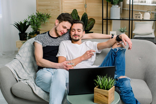 Portret gejowskiej pary relaksujący z białym winem na kanapie i oglądanie komputera. - Zdjęcie, obraz