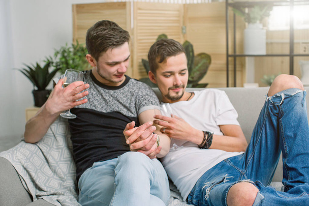 Young gay men holding hands on sofa at home. Shy gay couple showing love and romance, drinking wine - Фото, изображение