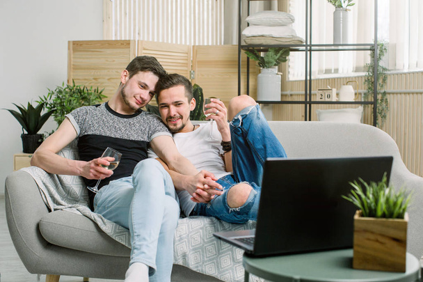 Young gay couple relaxing together with wine on a large comfortable sofa with a laptop computer surfing the internet, view from above - Foto, afbeelding