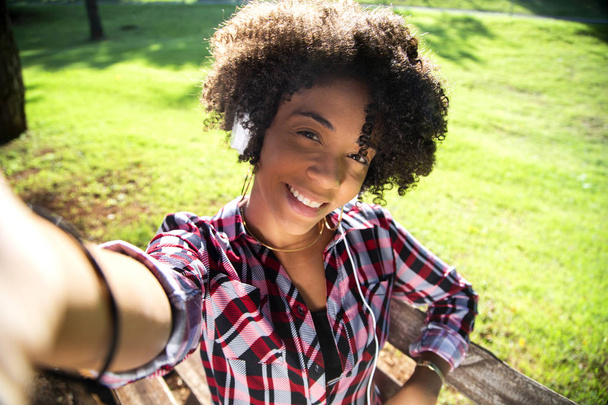 Primo piano ritratto di giovane donna afroamericana attraente sorridente in possesso di smartphone, scattare foto selfie e in piedi nel parco con piante sfocate sullo sfondo
  - Foto, immagini