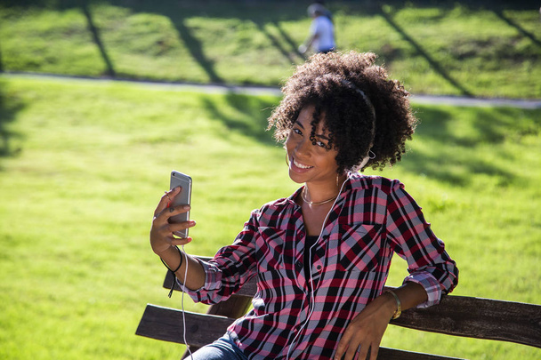 Closeup portrait of smiling young attractive African American woman holding smartphone, taking selfie photo and standing in park with blurred plants in background  - 写真・画像