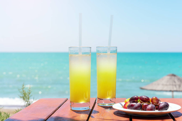 Two glasses of fresh tropical fruit juice on the beach, and a plate with grapes and watermelon on the table, against the sea. Holiday and vacation concept - Foto, immagini
