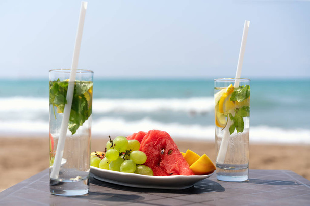 two glasses with lemon drink and ugly mint leaves on a table with a plate of fruit, on the beach, against the background of the sea - Foto, Bild