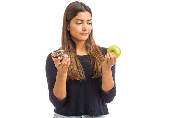 Hispanic confused woman looking at green apple and donut while standing against white background - Foto, Imagen