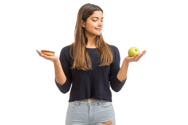 Confused woman in dilemma with donut and green apple against plain background - Photo, image