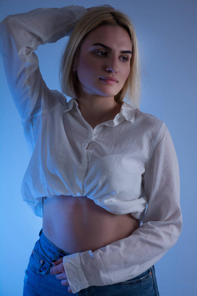 Fashion portrait of beautiful woman in white shirt with bare stomach and blue background in studio. Girl holding hand on head. Beauty and fashion concept - Fotó, kép