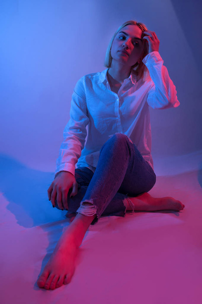 Όμορφο μοντέλο κορίτσι μόδας κάθεται στο στούντιο με πολύχρωμο μπλε και κόκκινο φως. Η γυναίκα φοράει άσπρο πουκάμισο, μπλε τζιν και με γυμνά πόδια. Χαμηλή ρύθμιση φωτισμού πλήκτρων - Φωτογραφία, εικόνα