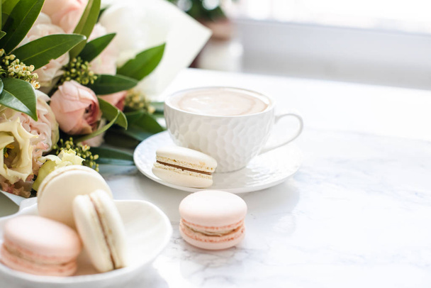 Tyylikäs makea jälkiruoka makaronit, kuppi kahvia ja pastellivärisiä beige kukkia kukkakimppu valkoinen marmori
 - Valokuva, kuva