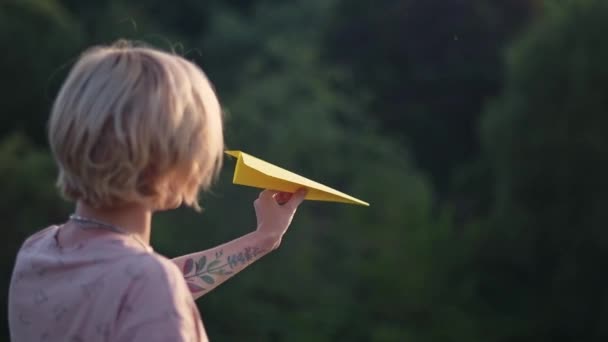 Een jong, mooi meisje met een tatoeage op haar hand lanceert een papieren vliegtuig. Mooie vrouw, buitenshuis in de natuur, bij zonsondergang, lanceert een gekleurd papier vliegtuig. - Video