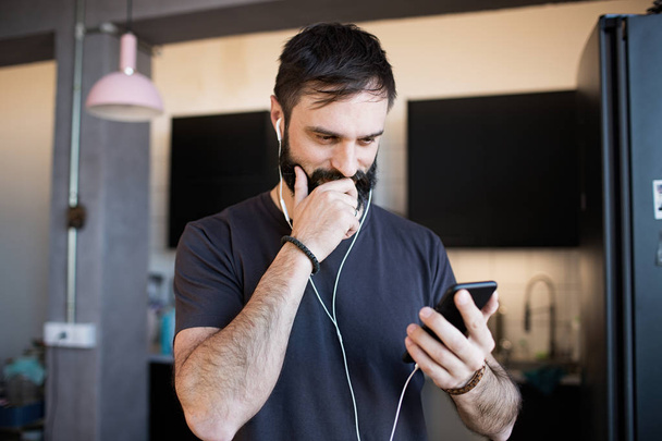 Handsome bearded guy wearing casual grey t-shirt listening music in earphones, checking social networks on smartphone having happy and delighted look. - Foto, Imagem