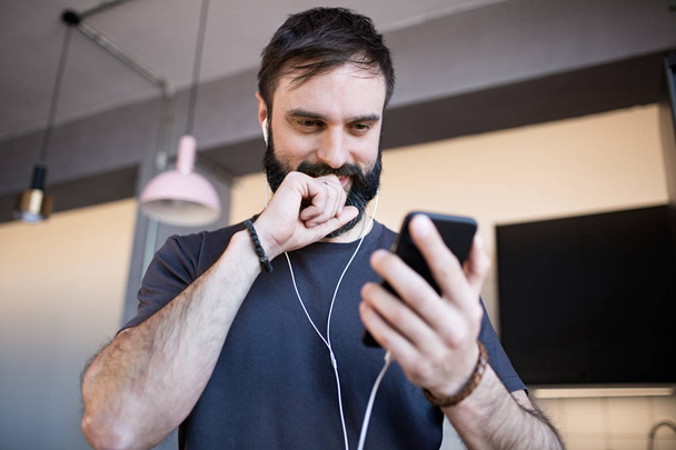 Handsome bearded guy wearing casual grey t-shirt listening music in earphones, checking social networks on smartphone having happy at home - Фото, зображення