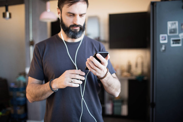 Trendy bearded guy wearing casual grey t-shirt listening music in earphones, checking social networks on smartphone having happy at home - Foto, Imagem