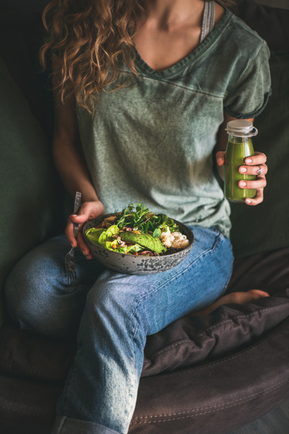 Здоровый ужин или обед. Женщина в футболке и джинсах сидит и держит веганский суперкубок или миску Будды с хумусом, овощами, салатом, бобами, кускусом и авокадо и смузи в руках
 - Фото, изображение
