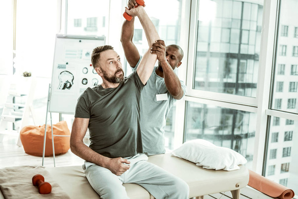 Careful bearded male person lifting dumbbell with help - 写真・画像