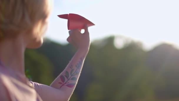 Een jong, mooi meisje met een tatoeage op haar hand lanceert een papieren vliegtuig. Mooie vrouw, buitenshuis in de natuur, bij zonsondergang, lanceert een gekleurd papier vliegtuig. - Video