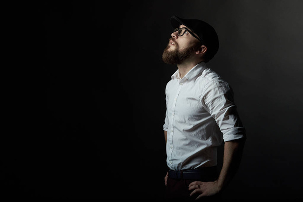 Portrait of a young man in glasses with a beard and a mustache in a black cap and a white shirt, A dark studio background - 写真・画像