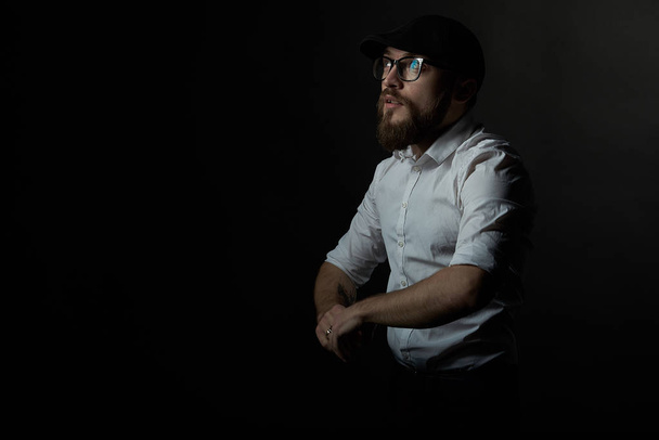 young handsome man in glasses with a beard and a mustache in a black cap and a white shirt holds his hands in front of him, a dark studio background  - Photo, Image