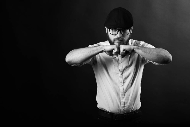 jeune homme beau dans des lunettes avec une barbe et une moustache dans une casquette noire et une chemise blanche tient ses mains devant lui, un fond sombre studio
  - Photo, image