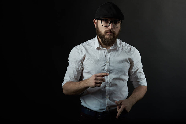 joven hombre guapo en gafas con una barba y un bigote en una gorra negra y una camisa blanca sostiene sus manos delante de él, un fondo oscuro estudio
  - Foto, imagen