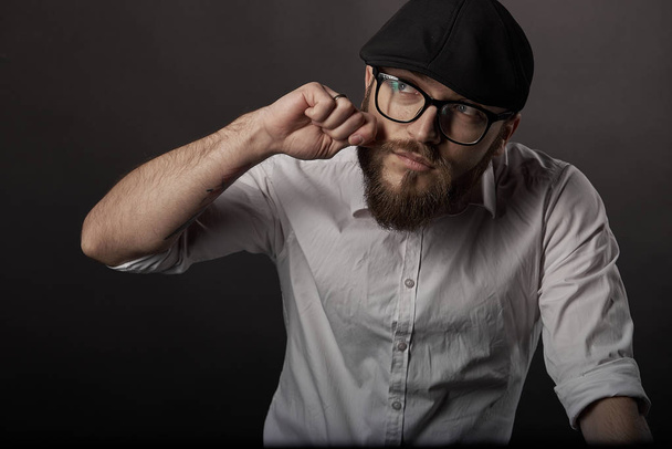 jonge knappe man met bril met een baard en een snor in een zwarte pet en een wit overhemd bevestigt zijn snor met zijn hand, de achtergrond van een donker studio  - Foto, afbeelding