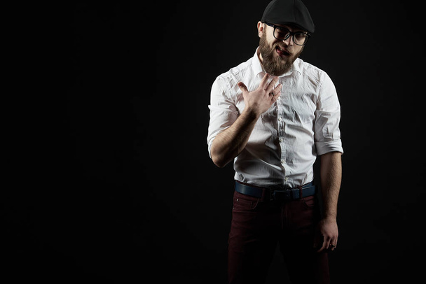 young handsome man in glasses with a beard and a mustache in a black cap and a white shirt shows a variety of hand gestures, a dark studio background - Photo, Image