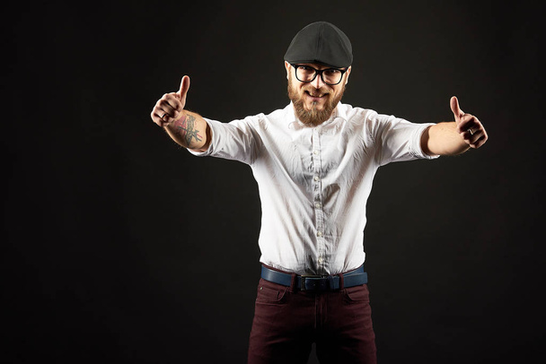 young handsome man in glasses with a beard and a mustache in a black cap and a white shirt shows a variety of hand gestures, a dark studio background - Photo, Image