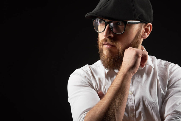 Portrait of a young man in glasses with a beard and a mustache in a black cap and a white shirt, A dark studio background - Photo, Image