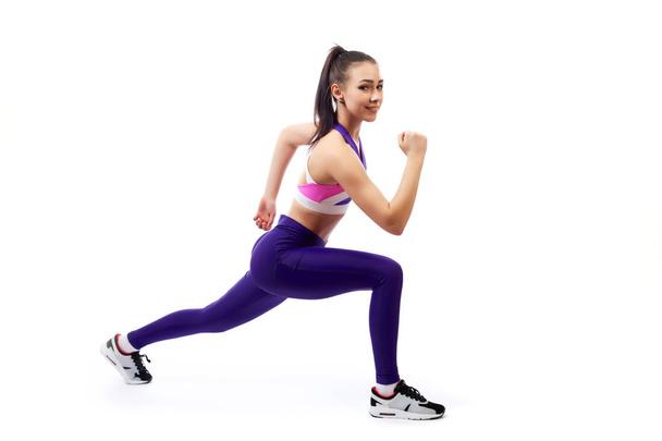 A young  woman coach in a sporty   short top and gym leggings makes lunges  by the feet forward, hands are held out to the side   on a  white isolated background in studio  - Photo, Image