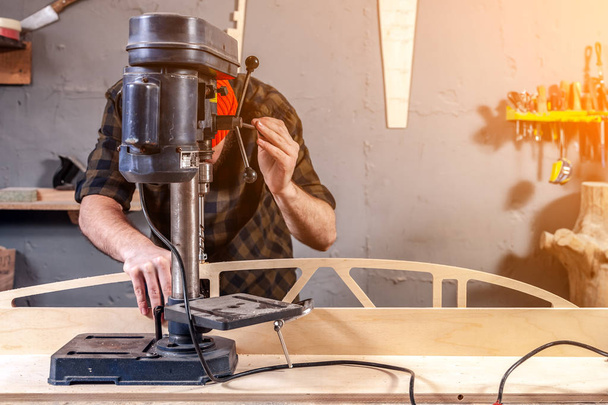 Primer plano de un hombre con ropa de trabajo y una gorra de carpintero está tallando una tabla de madera en una gran máquina de perforación moderna en un taller de luz. Conceptos de reparación
  - Foto, imagen