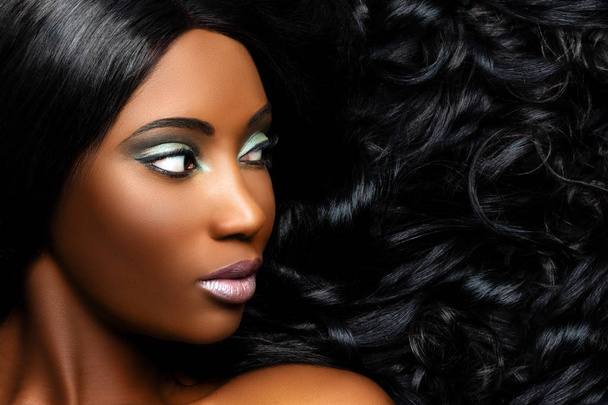 Extremo primer plano retrato de belleza de hermosa joven africana con maquillaje profesional. Chica mirando a un lado con el pelo largo rizado al lado de la cara
. - Foto, imagen