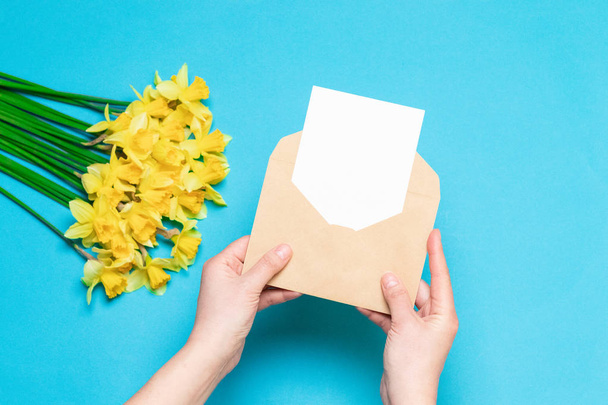 Office desk, spring concept. Women's hands hold craft envelope with a postcard, fresh yellow daffodils on the background - Photo, Image