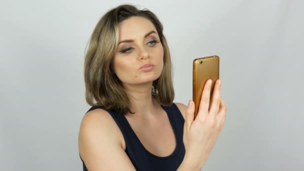 Portrait d'une belle jeune femme qui pose faisant un selfie et communiquant et regardant dans un smartphone qui tient dans ses mains sur fond blanc en studio
 - Séquence, vidéo