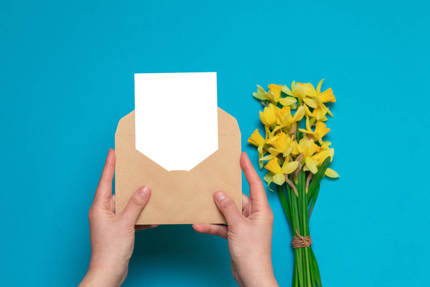manos femeninas sosteniendo sobre artesanal con una tarjeta, narcisos amarillos frescos sobre un fondo azul. Escritorio de oficina, concepto de primavera
. - Foto, imagen