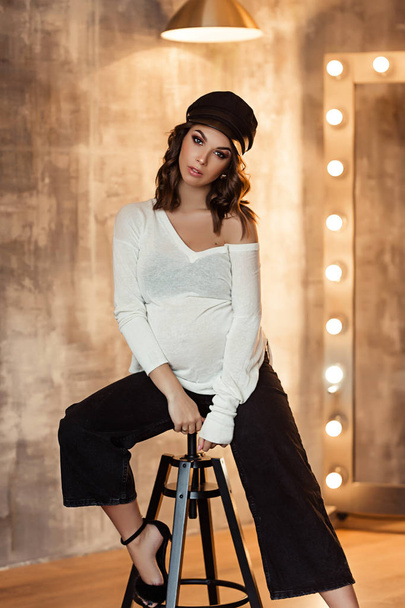Pregnant european woman in white blouse and black pants with a black hat in dark flat, stylish and happy pregnant woman in apartments, beautiful pregnant woman with dark hair - Foto, Bild