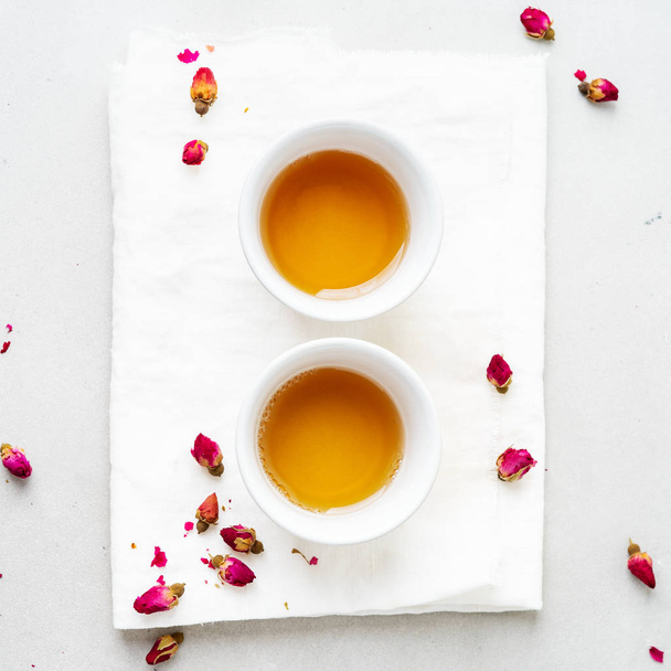 Two white ceramic cups of herbal tea with dried pink rose buds on gray background  - Фото, зображення