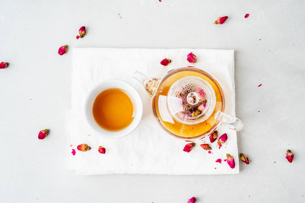 white ceramic cup and transparent teapot with herbal tea of dried pink roses on gray background - Fotografie, Obrázek