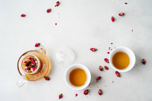 two white cups and transparent teapot with herbal tea of dried pink roses buds on gray stone background - Photo, Image