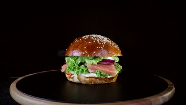 Fast Food. Hamburger met rode vis, zalm en forel greens, tomaat en ui, langzaam spinnen op een houten bord op een zwarte achtergrond - Video