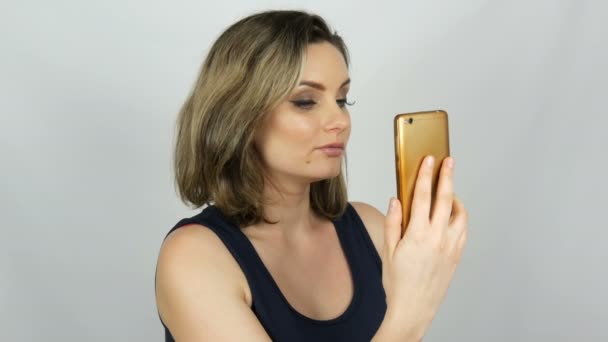 Portret van een mooie jonge vrouw die poseert het maken van een selfie en communiceren en op zoek naar een smartphone die in haar handen houdt op witte achtergrond in de Studio - Video