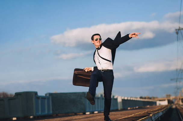 Young Belarusian businessman in a suit with a suitcase rejoices in successes in his career. Good news. Downshifting, vacation concept. - 写真・画像