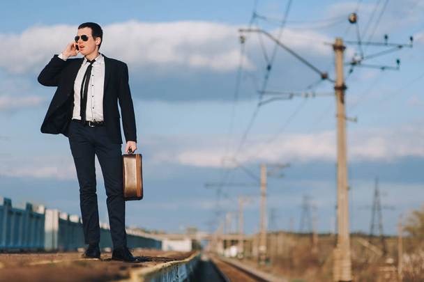 Handsome young business man in sunglasses standing on a railroad station with his suitcase and calling by phone. Evening blue sky with clouds. - Фото, изображение