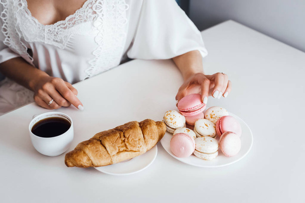 Primer plano de la mujer en camisón blanco sosteniendo pasteles macaron mientras el desayuno
 - Foto, imagen