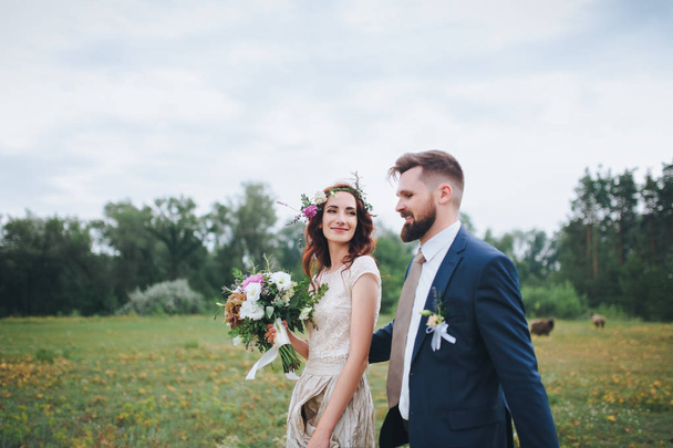 The bride and groom walking  along the forest glade leaning against each other. The bride with a wreath of fresh flowers and with a bouquet. - Φωτογραφία, εικόνα