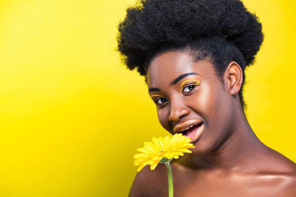atemberaubende afrikanisch-amerikanische Frau mit Blume schaut in die Kamera auf gelb  - Foto, Bild